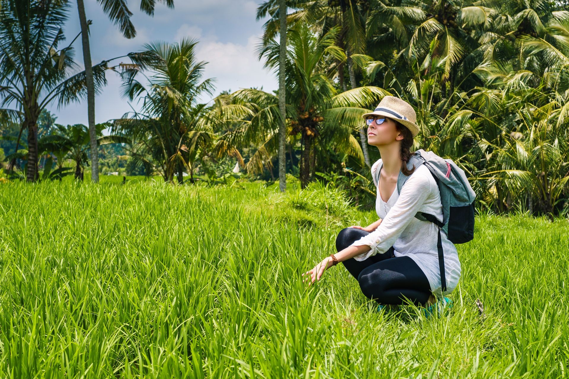 Beautiful young girl traveler in hat and with backpack on the rice field, in high green grass, with palms, amazing blue sky with white clouds on the background has journey in Bali island Indonesia